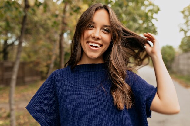 Happy smiling lady with wavy hair wearing blue sweater posing touching her hair in the park