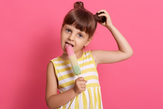 水の氷をなめるとピンクに分離されたポーズ彼女の面白い結び目に触れながら幸せな小さな子供の笑顔