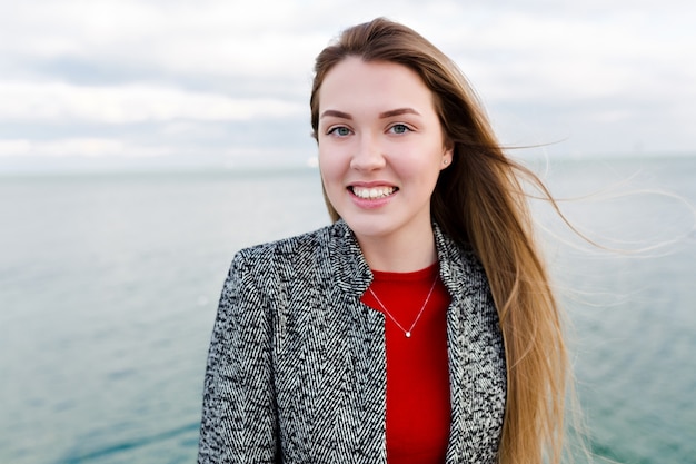 Happy smiling long-haired woman with big blue eyes in red shirt and grey coat walks near the sea