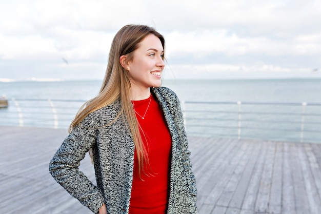 Happy smiling long-haired woman with big blue eyes in red shirt and grey coat walks near the sea