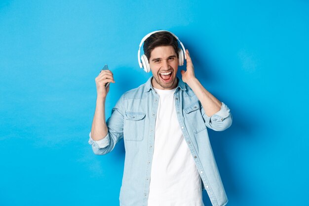 Счастливый улыбающийся человек наслаждается прослушиванием музыки в наушниках, держа смартфон в поднятой руке, стоя на синем фоне
