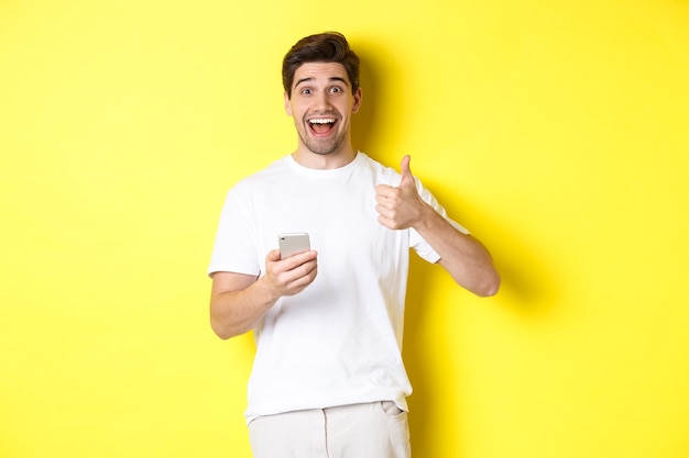 Happy smiling man holding smartphone, showing thumb up in approval, recommend something online, standing over yellow background.