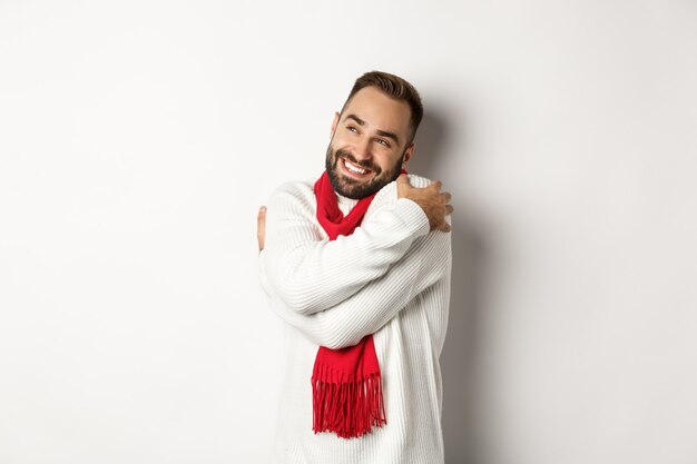 Happy smiling man hugging himself and looking satisfied at upper left corner, wearing warm and comfortable winter sweater, standing over white background.
