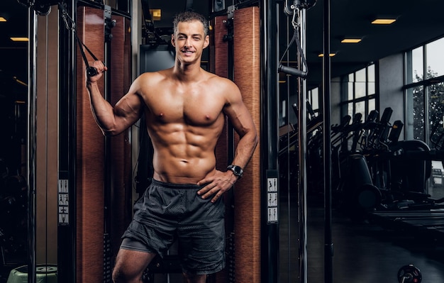 Happy smiling man is doing exercises with training apparatus at dark gym club.