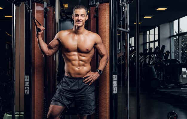 Happy smiling man is doing exercises with training apparatus at dark gym club.