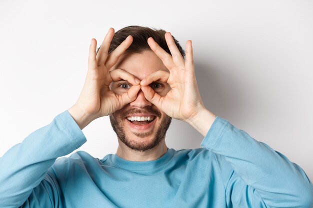 Happy smiling man looking through hand binoculars with amazed face, checking out promo offer, standing on white background.