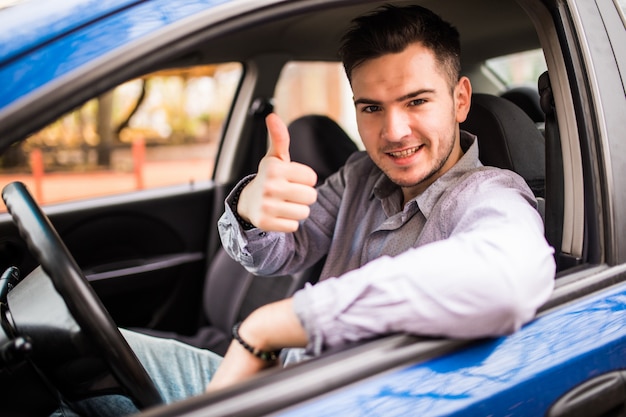 Счастливый улыбающийся человек, сидящий внутри автомобиля, показывает палец вверх. Красивый парень в восторге от своего нового автомобиля. Положительное выражение лица