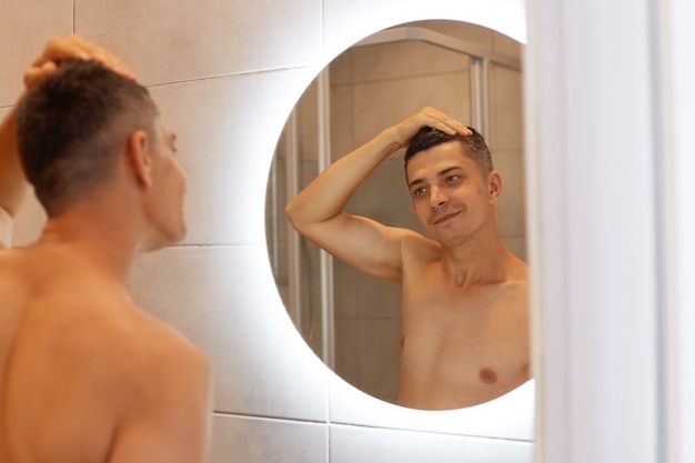 Happy smiling man standing with naked upper body and looking smiling at his Reflection in the mirror, posing in bathroom after taking shower in morning.