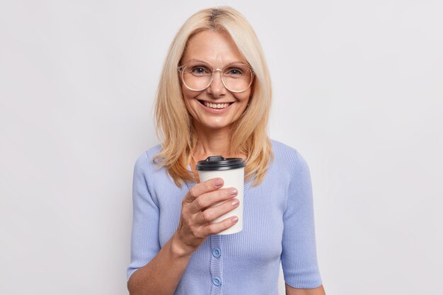 Happy smiling mature woman with blonde hair buys caffeine drink in cafe holds disposable cup of coffee wears transparent glasses casual blue jumper isolated over white wall. Lifestyle.