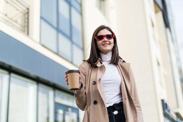 Happy smiling model in light brown coat drinks coffee outside
