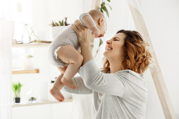 Happy smiling mother playing with newborn child in comfy light bedroom in front of window. Moments of motherhood happiness with kids. Family concept.