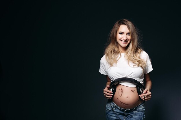 Happy and smiling pregnant female wearing white t shirt and jeans with big earphones on tummy Expectant mother painting funny smile on her naked belly Concept of maternety and health care