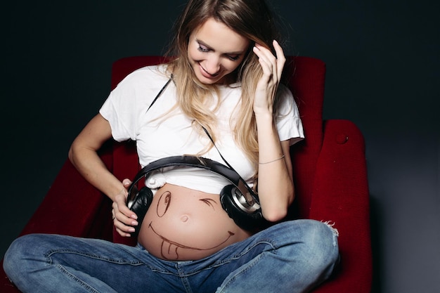 흰색 티셔츠와 청바지를 입고 배에 큰 이어폰을 꽂은 행복하고 웃는 임산부 임산부는 벌거벗은 배에 재미있는 미소를 그린다 출산과 건강 관리의 개념