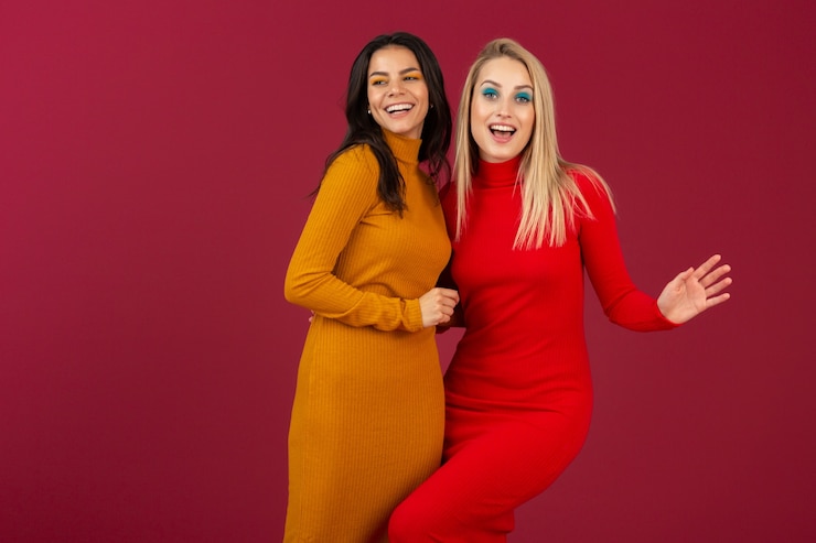 Free Photo happy smiling pretty stylish women in yellow and red autumn winter fashion knitted dress posing isolated on red wall