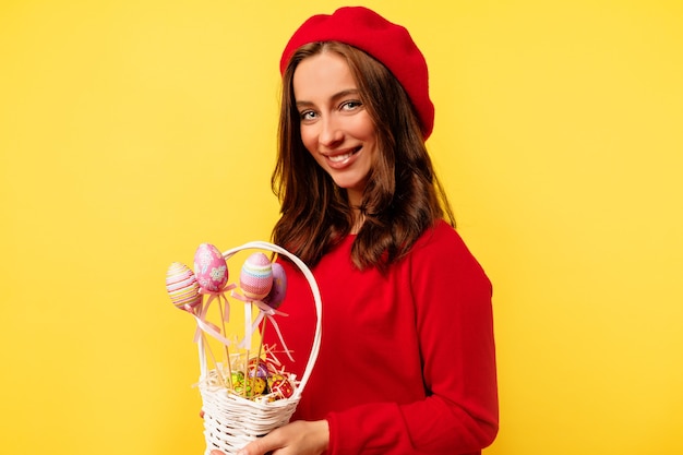 孤立した黄色の壁の上にイースターバスケットでポーズをとって赤いプルオーバーと赤いベレー帽を身に着けている巻き毛の幸せな笑顔のきれいな女性