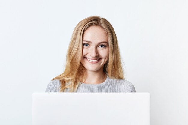 Happy smiling self-employed woman works at home, sits in front of opened laptop, enjoys free wireless internet connection. Lovely teenage girl messages with friends, has pleasant smile on face