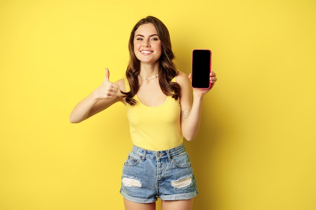 Ragazza alla moda sorridente felice che mostra i pollici in su e l'interfaccia dell'app per smartphone, il display del telefono cellulare, soddisfatta del sito web, dell'applicazione per lo shopping online, in piedi su sfondo giallo.