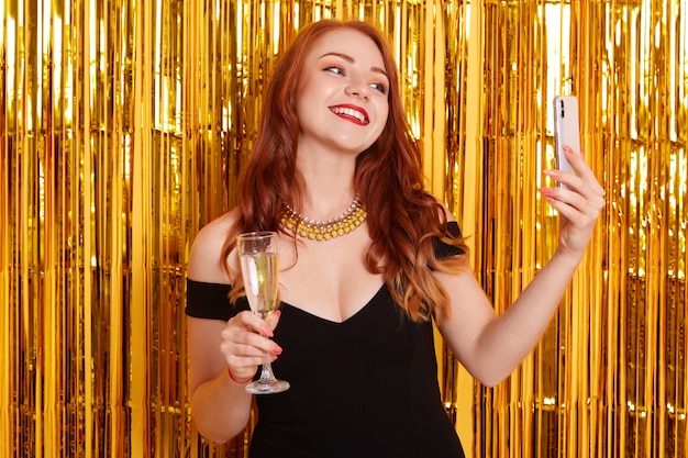 Sorridere felice con fare posa contro orpelli d'oro e fare selfie tramite un moderno smart phone, ragazza che indossa un abito nero, signora che tiene un bicchiere di vino, che celebra un evento importante.