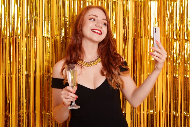 Happy smiling with make posing against golden tinsel and making selfie via modern smart phone, girl wearing black dress, lady holding glass of wine, celebrating important event.