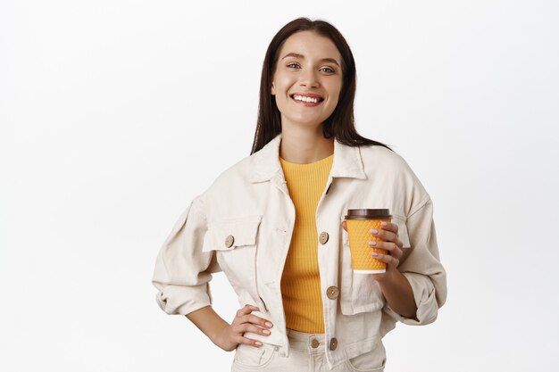 Счастливая улыбающаяся женщина пьет кофе из желтой чашки на вынос, покупает напиток в кафе и гуляет по городу, довольная стоя на белом