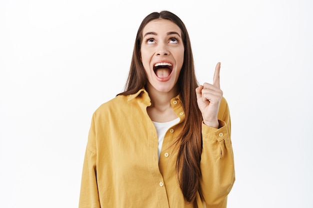 Happy smiling woman gasping astonished, looking in awe above, pointing finger up at cool new promo deal, shopping offer discount, standing against white wall