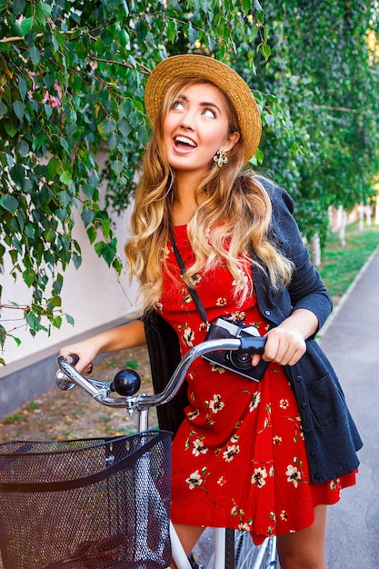 Happy smiling woman having fun and surprised playful emotions, walking alone with stylish retro bike in city park, wearing red dress warm sweater and vintage straw hat, have curled long blonde hairs.