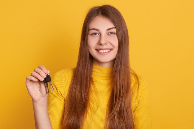 Happy smiling woman holding key in hands, wearing casual shirt, having long beautiful hair, buys new flat, looks happy, expressing positive emotions, being lucky.