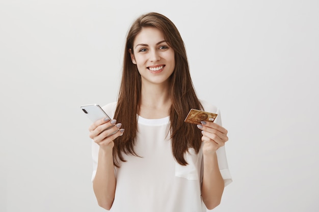 Happy smiling woman order online via smartphone app, holding credit card and mobile phone