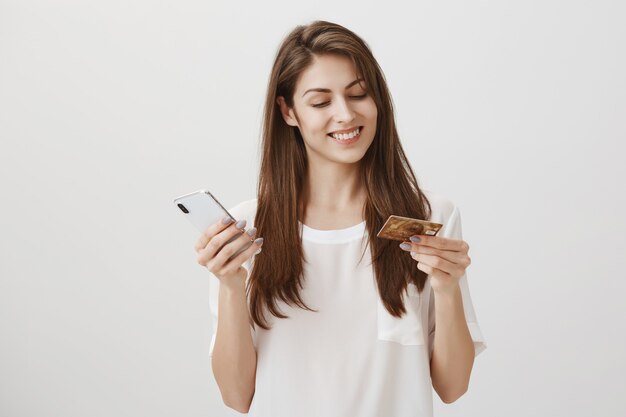 Happy smiling woman order online via smartphone app, looking at credit card