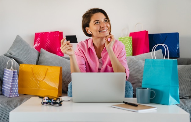 Счастливая улыбающаяся женщина в розовой рубашке на диване у себя дома среди красочных сумок с кредитной картой, оплачивающей онлайн на ноутбуке