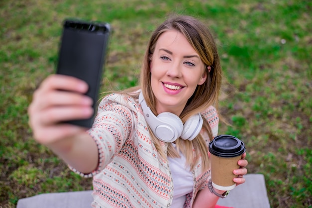 Happy smiling woman sitting on grass and makes self-portrait on smartphone. Woman Hand holding Paper Cup coffee Outdoor. Portrait of smiling woman in the park and making funny selfie, looking at camera.