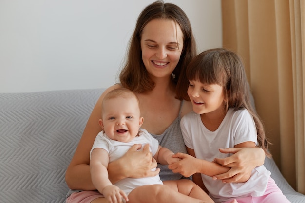 Happy smiling woman sitting with her children on sofa, family spending time indoor at home, laughing happily, talking, people wearing casual style clothing.