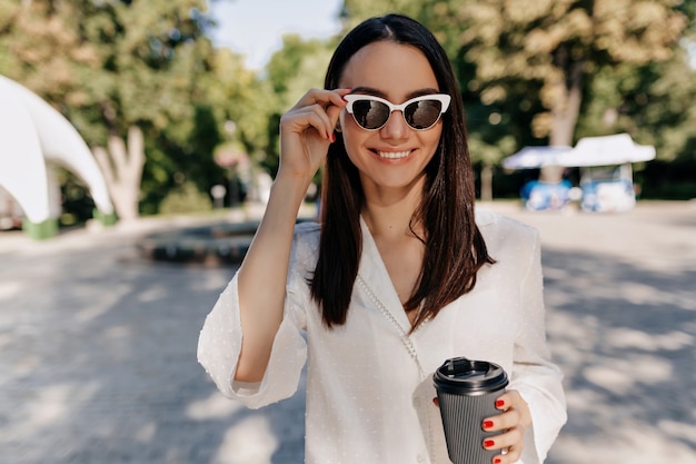 Happy smiling woman wearing white shirt and white glasses drinking coffee outside in good sunny day in the city park