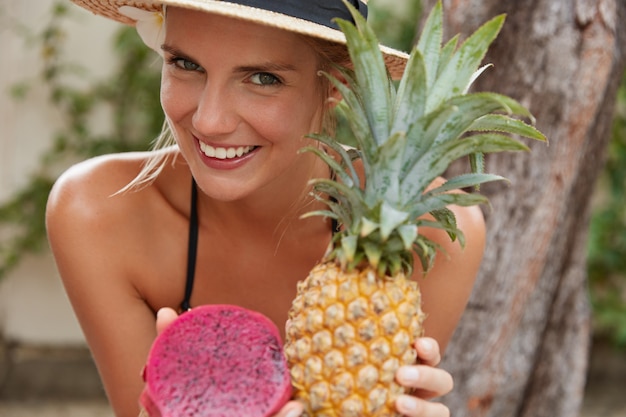 Happy smiling woman with healthy skin, has broad smile, eats exotic fruits, has good recreation in tropical country, spends summer vacation in paradise place, recieves vitamins. Healthy eating