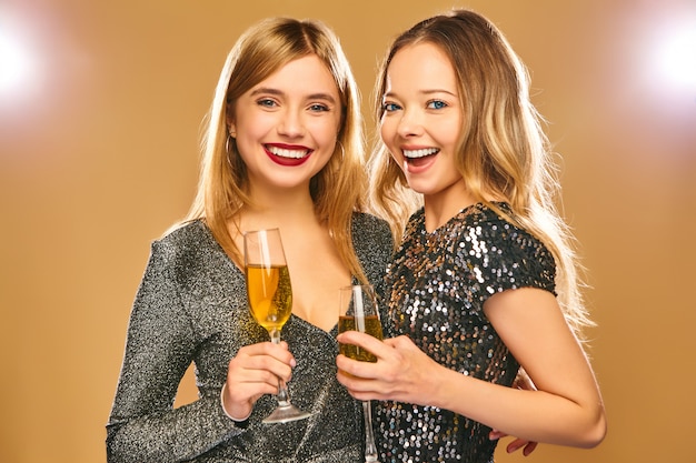 Счастливые улыбающиеся женщины в стильных гламурных платьях с бокалами шампанского на золотой стене