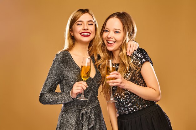 Счастливые улыбающиеся женщины в стильных гламурных платьях с бокалами шампанского на золотой стене