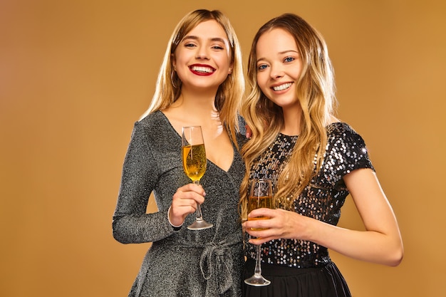 Happy smiling women in stylish glamorous dresses with champagne glasses on golden wall