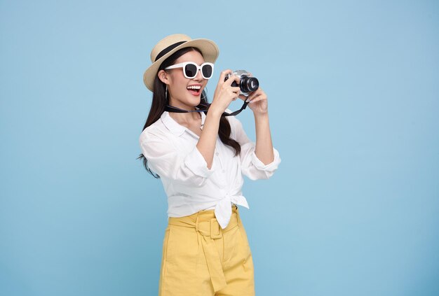 Счастливая улыбающаяся молодая азиатская женщина-туристка в летней шляпе, стоящая с камерой и фотографирующая
