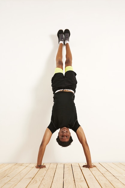 happy smiling young black model in black and yellow workout clothes doing handstand against a white wall on wooden floor.