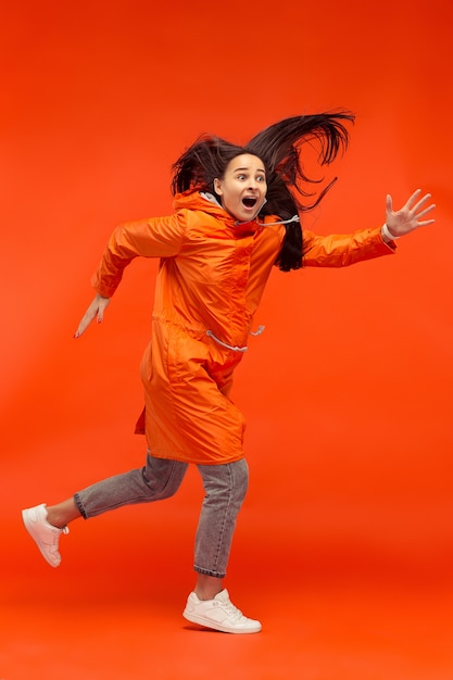 Счастливая улыбающаяся молодая девушка позирует в студии в осенней оранжевой куртке, изолированной на красном.