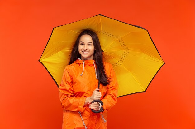 The happy smiling young girl posing at studio in autumn orange jacket isolated on red.