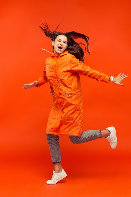 The happy smiling young girl posing at studio in autumn orange jacket isolated on red.
