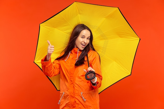 The happy smiling young girl posing at studio in autumn orange jacket isolated on red.