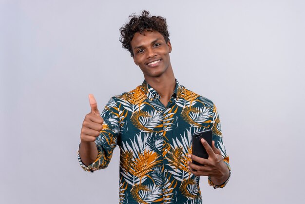 Happy and smiling young handsome dark-skinned man with curly hair in leaves printed shirt holding mobile phone while posing with thumbs up sign 