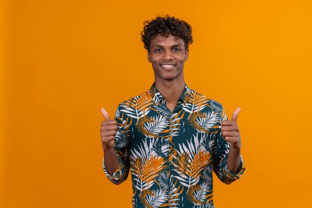 Счастливый и улыбающийся молодой красивый темнокожий мужчина с вьющимися волосами в рубашке с принтом листьев показывает палец вверх