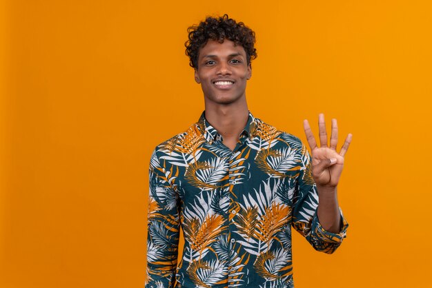 Счастливый и улыбающийся молодой красивый темнокожий мужчина с вьющимися волосами в рубашке с принтом листьев, показывая пальцами номер четыре
