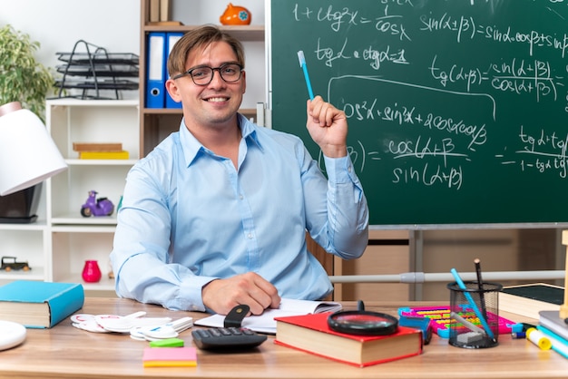 Happy and smiling young male teacher sitting at school desk with books and notes holding pencil in front of blackboard in classroom