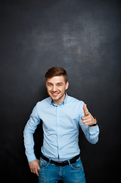 happy smiling young man in blue shirt over blackboard point with a finger in camera