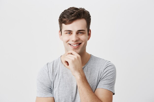 행복 하 게 웃는 젊은 남자 긍정적 인 감정 또는 감정을 보여줍니다, 유행 머리를 부담없이 입고, 턱에 손을 유지, 텍스트 또는 광고 복사 공간 회색 벽에 서.