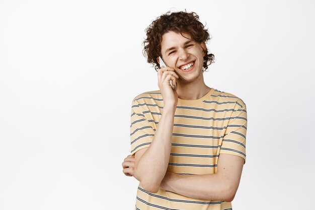 Happy smiling young man talking on mobile phone laughing over a call standing in tshirt over white background